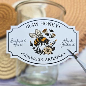 May include: White label with black text and border, reading "RAW HONEY", "Backyard Hives", "Hand Gathered", and "Surprise, Arizona". A bee with yellow and black stripes is illustrated in the center, surrounded by flowers.