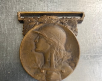 May include: A bronze medal featuring a profile of a woman wearing a helmet and laurel wreath. The medal has a loop at the top for attaching to a ribbon.