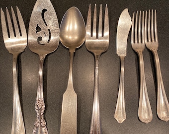 May include: A set of eight vintage silver-plated utensils, including a serving spoon, a cake server, a butter knife, and five forks. The utensils have ornate handles and are in good condition.