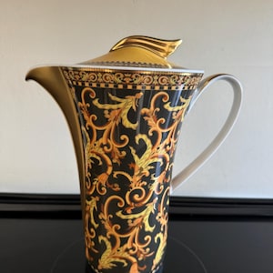 Pode incluir: Uma chaleira de cerâmica preta e dourada com uma alça e tampa decorativas. A chaleira apresenta uma borda dourada e um padrão barroco detalhado em ouro e laranja. A alça é branca e a tampa tem um detalhe dourado.