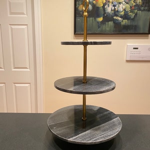 May include: A three-tiered serving stand made of black marble with a gold metal connecting rod. The stand has a round top and is perfect for displaying food or desserts.