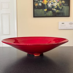 May include: A vibrant red ceramic bowl with a wide, shallow design. The bowl has a glossy finish and rests on three small, unglazed feet. The bowl's shape is modern and elegant, suitable for serving or display.