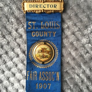 May include: A blue and gold ribbon with the text "Director", "St. Louis County", "Fair Assoc'n", and "1907". The ribbon has a gold circular pin with a picture of a farm scene.