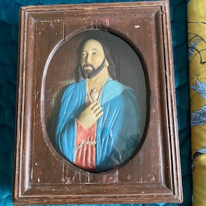 Puede incluir: Un relieve religioso enmarcado que representa una figura con pelo largo y barba, vestida con una túnica azul. La talla está dentro de un marco de madera rectangular marrón oscuro, con desgaste visible.