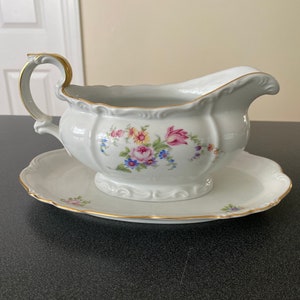 May include: White porcelain gravy boat with a floral pattern and gold trim. The boat is on a matching saucer.