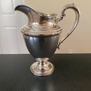May include: A silver pitcher with a rounded body and a flared spout. The pitcher has a decorative handle and sits on a pedestal base.