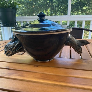 Può includere: Una ciotola da portata in ceramica nera con un coperchio e due manici a forma di pesce. La ciotola ha una finitura lucida e un bordo bianco.