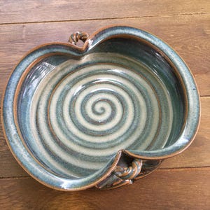 Op de afbeelding: Een blauwe en bruine keramische serveerschaal met een spiraalvormig ontwerp en twee handvatten. De schaal is handgemaakt en heeft een rustieke uitstraling.