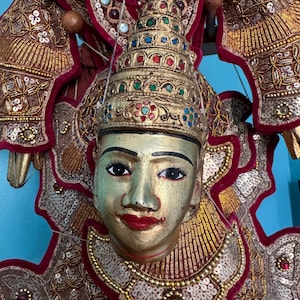 Puede incluir: Un títere birmano tradicional con una cara pintada y un tocado elaborado adornado con oro y joyas. El títere lleva un traje rojo y dorado con lentejuelas y cuentas.