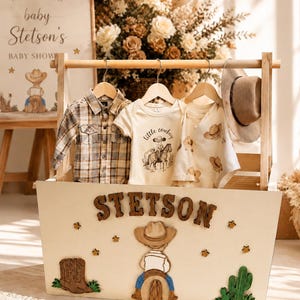 May include: A wooden baby clothes rack containing a plaid shirt, a cream-colored bodysuit with the text little cowboy, and a patterned bodysuit. A tan hat hangs on the side. The front of the rack features the name STETSON, stars, boots, a cowboy, and a cactus.