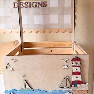 May include: Wooden toy box with a nautical theme. Features a lighthouse, sailboats, seagulls, and waves. The box has a handle and the text "TB DESIGNS" on the top. The box is made of light-colored wood.