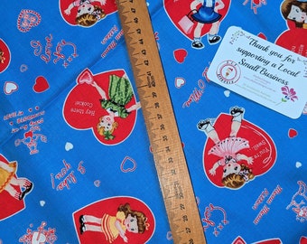 May include: Blue fabric with a repeating pattern of red hearts featuring cartoon girls in vintage dresses. The text "Love... You" and "Hey there Cookie!" are printed on the fabric. A wooden ruler is placed on top of the fabric.