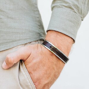 May include: A brown leather bracelet with a silver band and engraved coordinates. The bracelet is worn on a person's wrist.
