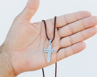 May include: A silver cross pendant necklace with a brown cord. The cross has an arrow design and the letters "GMV". The necklace is held against a white background.