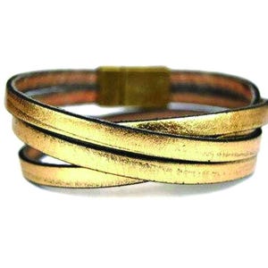 May include: A gold leather wrap bracelet with a gold metal clasp. The bracelet is made of multiple strands of leather that are woven together.