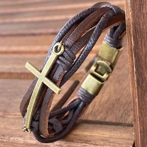May include: A brown leather bracelet with a woven design and a gold-toned cross charm.