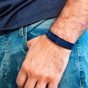 May include: A blue and black bracelet with a smooth, flat surface. The bracelet is worn on the wrist of a person wearing blue jeans.