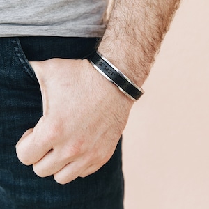 May include: A black leather bracelet with a silver band engraved with the coordinates "46.58.21.N 71.31.17.W".