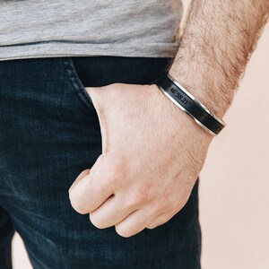 May include: A black leather cuff bracelet with a silver band engraved with the coordinates "46.58.21.N 119.37.57.W".