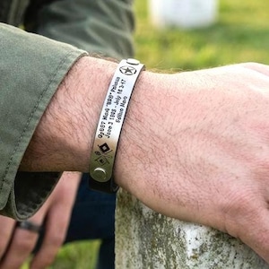 Könnte beinhalten: Ein metallisches Armband mit einer Gravur, die "GySgt Mark "BBQ" Pelossa June 2 1983 - July 16 17" lautet, sowie einem Sternsymbol und einer Rautenform. Das Armband hat ein dunkles Band und eine silberfarbene Metallplatte mit sichtbaren Nieten.