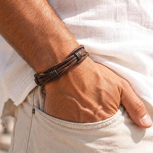 Puede incluir: Una pulsera de cuero marrón oscuro compuesta por varias tiras finas superpuestas y aseguradas con pequeños anillos de metal oscuro. La pulsera se lleva en la muñeca, combinada con una camisa de lino blanca y pantalones claros en un entorno exterior.