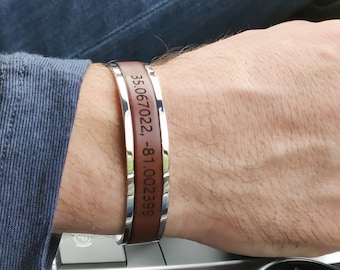 May include: A cuff bracelet featuring a polished silver-tone metal frame and a central brown band. The brown band is engraved with the black text 35.067022, -81.002399. The bracelet has a rigid, open-ended design that wraps around the wrist.