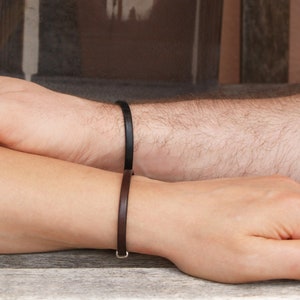 May include: Two wrists with matching leather bracelets. The bracelet on the left wrist is brown, and the bracelet on the right wrist is black.