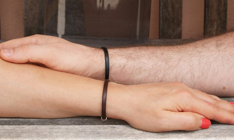 May include: Two wrists with matching leather bracelets. The bracelet on the left wrist is brown, and the bracelet on the right wrist is black.