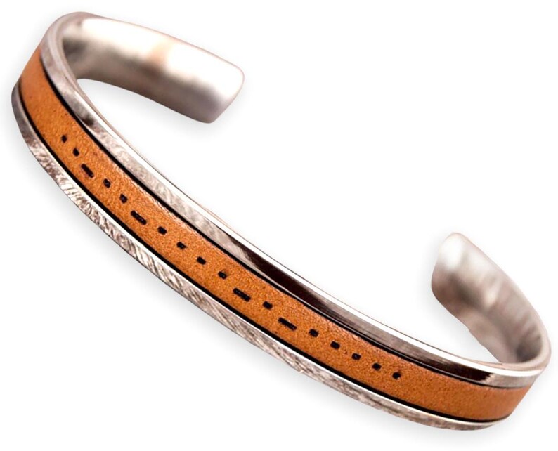 K&ouml;nnte beinhalten: Ein silbernes Armreif-Armband mit einem hellbraunen Lederband. Das Lederband ist mit schwarzem Faden gen&auml;ht. Das Armband hat eine strukturierte Silberoberfl&auml;che an den Seiten.