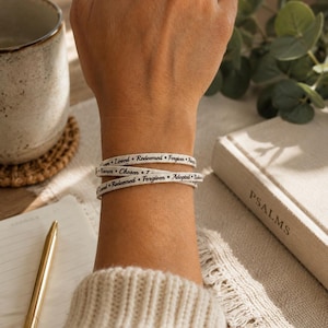 Puede incluir: Una pulsera envolvente blanca con texto impreso en negro que incluye las palabras Loved, Redeemed, Forgiven, Adopted, Known y Chosen, separadas por peque&ntilde;os puntos negros. La pulsera se lleva alrededor de la mu&ntilde;eca en estilo multicapa.
