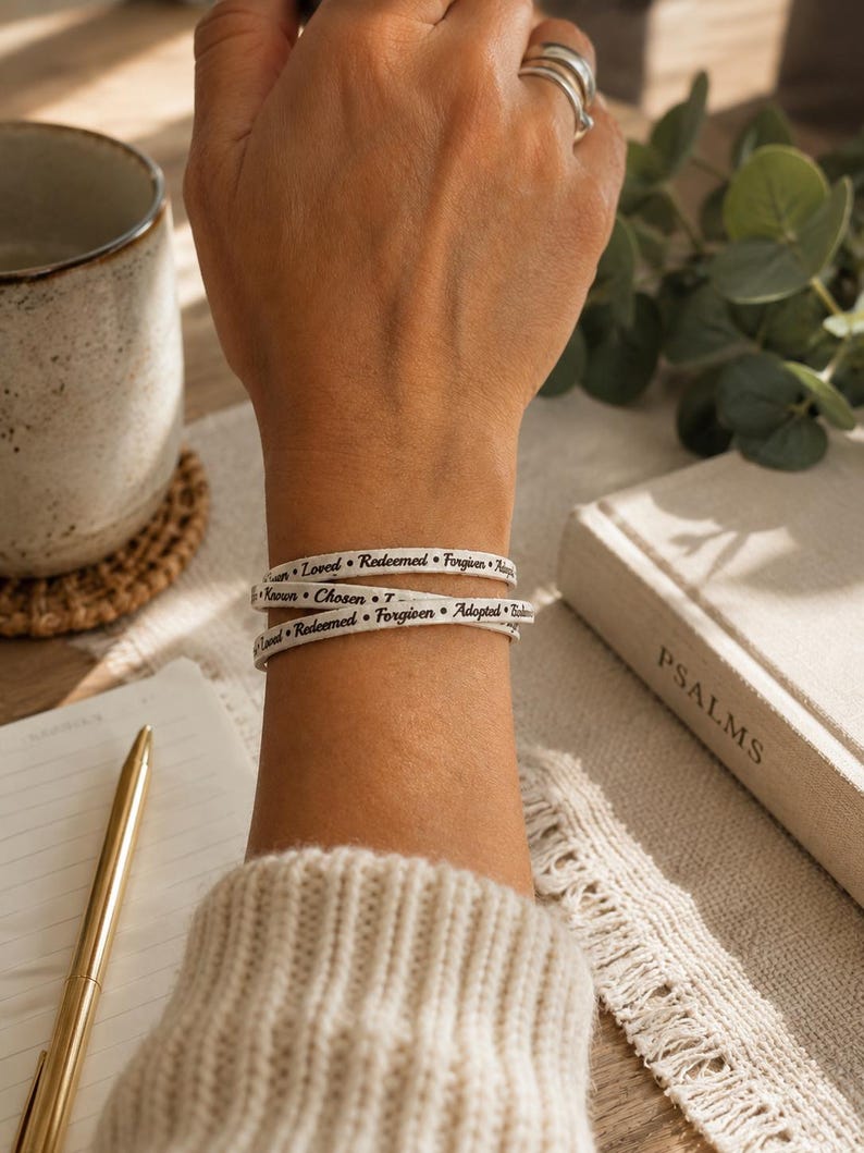 Puede incluir: Una pulsera envolvente blanca con texto impreso en negro que incluye las palabras Loved, Redeemed, Forgiven, Adopted, Known y Chosen, separadas por peque&ntilde;os puntos negros. La pulsera se lleva alrededor de la mu&ntilde;eca en estilo multicapa.