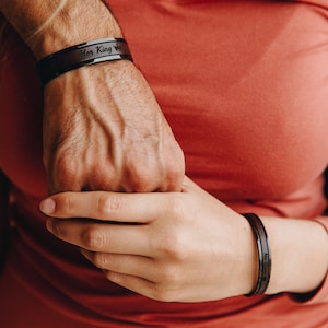 May include: A close-up of two hands holding each other. The person on the left is wearing a black bracelet with the words "Her King" engraved on it. The person on the right is wearing a black bracelet.