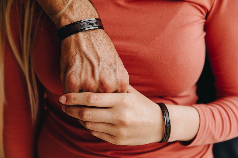 May include: A close-up of two hands holding each other. The person on the left is wearing a black bracelet with the words "Her King" engraved on it. The person on the right is wearing a black bracelet.