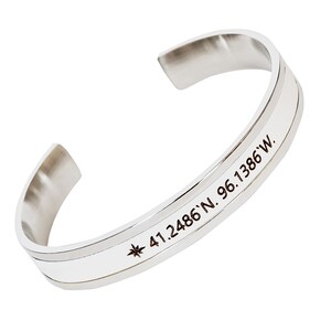 May include: Silver bangle bracelet with a compass and coordinates engraved on it. The coordinates are 41.2486'N 96.1386'W.