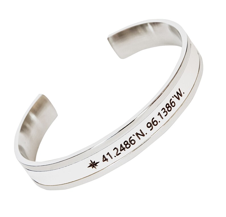 May include: Silver bangle bracelet with a compass and coordinates engraved on it. The coordinates are 41.2486'N 96.1386'W.