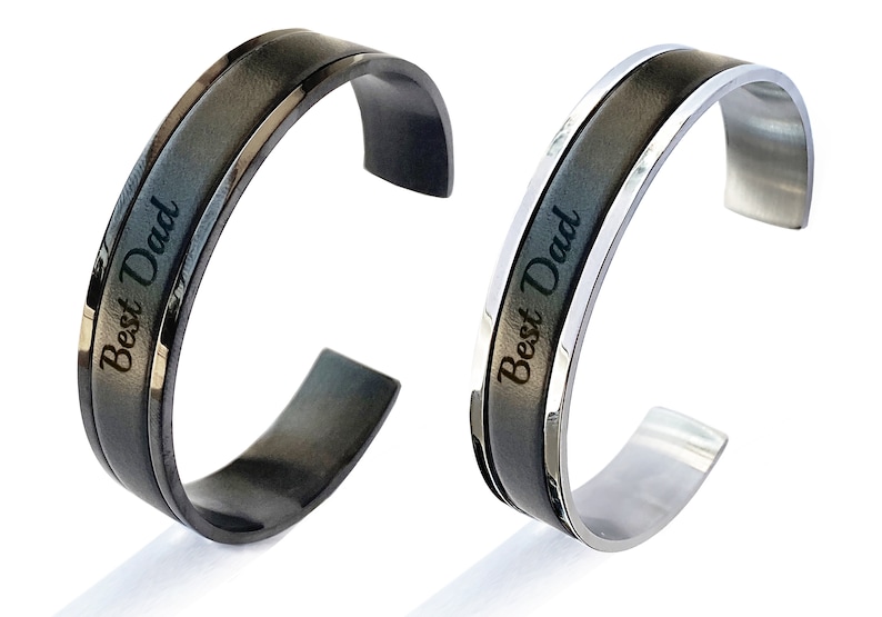 May include: Two open cuff bracelets. One is black with a dark grey centre band, the other is silver with a dark grey centre band. Both have the words "Best Dad" engraved on the centre band.