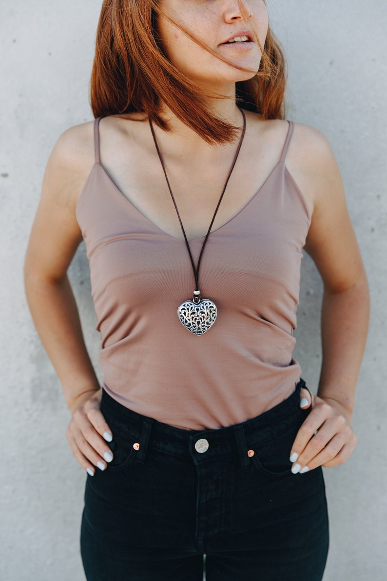 May include: A woman wearing a light brown tank top with a silver heart pendant necklace. The pendant has a delicate, intricate design. She is also wearing black jeans.