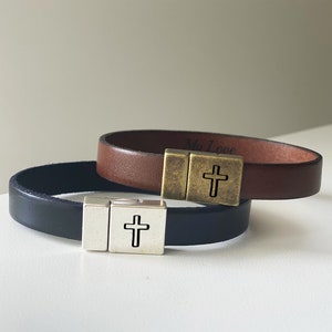 Puede incluir: Dos pulseras de cuero con un dije de cruz. La pulsera de arriba es de cuero marrón con las palabras "My Love" grabadas en el interior. La pulsera de abajo es de cuero negro.