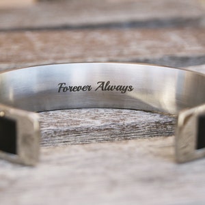 May include: A silver cuff bracelet with a black leather band and the inscription "Forever Always" engraved on the inside.
