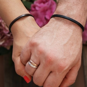 May include: Two people holding hands, each wearing a simple black leather bracelet. The woman is wearing a ring on her finger.