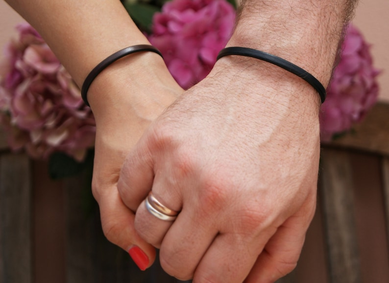 May include: Two people holding hands, each wearing a simple black leather bracelet. The woman is wearing a ring on her finger.