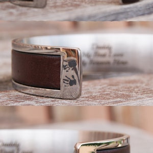 May include: A silver cuff bracelet with a brown leather band. The bracelet is engraved with the words "Daddy, My First Love, My Forever Hero." The leather band has a small imprint of two footprints.