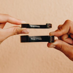 May include: Two black metal bracelets with brown leather accents. Each bracelet has the words "Always & Forever" engraved on the front.