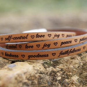 Puede incluir: Un brazalete de cuero marr&oacute;n con las palabras "love", "joy", "peace", "patience", "kindness", "goodness", "faithfulness" y "self-control" grabadas en &eacute;l. Cada palabra est&aacute; separada por un s&iacute;mbolo de coraz&oacute;n.
