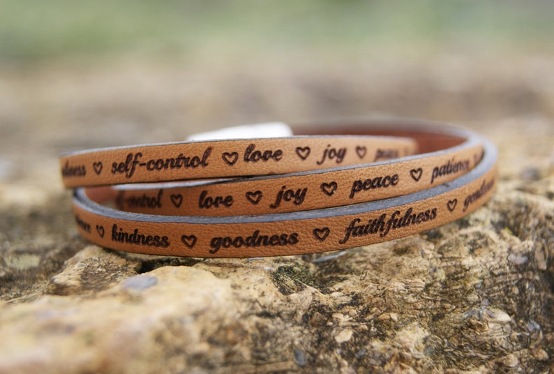 Puede incluir: Un brazalete de cuero marr&oacute;n con las palabras "love", "joy", "peace", "patience", "kindness", "goodness", "faithfulness" y "self-control" grabadas en &eacute;l. Cada palabra est&aacute; separada por un s&iacute;mbolo de coraz&oacute;n.