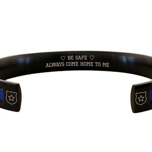 May include: Black stainless steel bracelet with a blue line and a star engraved on the side. The bracelet has an inscription that reads "BE SAFE &hearts; ALWAYS COME HOME TO ME".