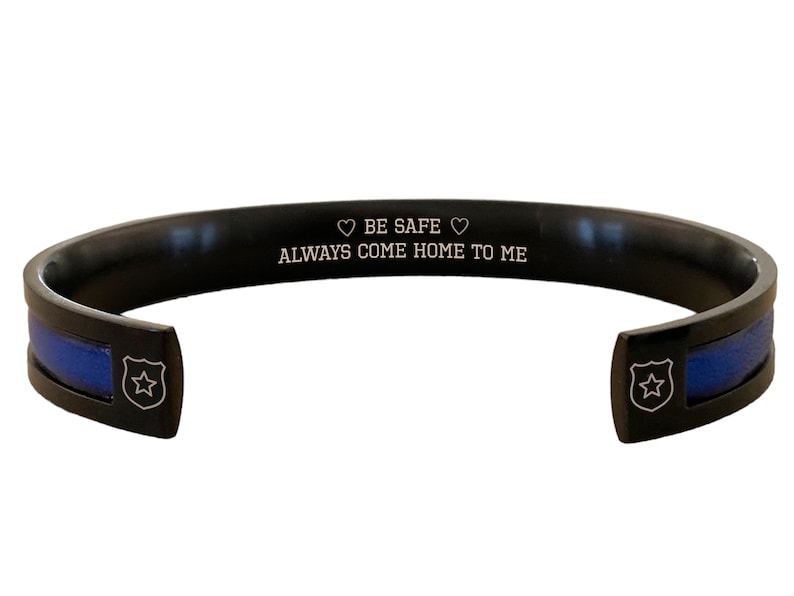 May include: Black stainless steel bracelet with a blue line and a star engraved on the side. The bracelet has an inscription that reads "BE SAFE &hearts; ALWAYS COME HOME TO ME".