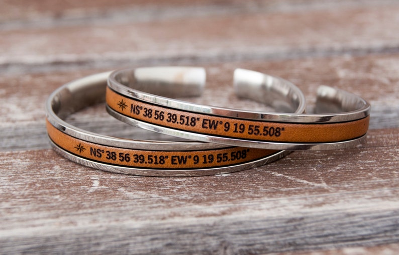 May include: Two silver bracelets with brown leather bands. Each bracelet has a compass symbol and engraved coordinates: *NS&deg; 38 56 39.518" EW9 19 55.508"