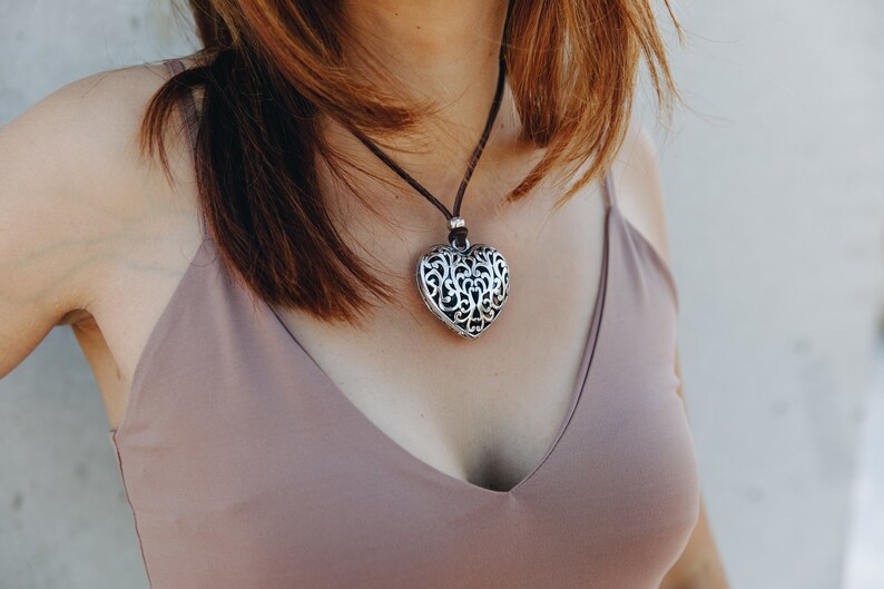 May include: A silver heart-shaped pendant necklace with a delicate filigree design hangs on a brown leather cord. The necklace is worn by a person with brown hair and a light brown tank top.