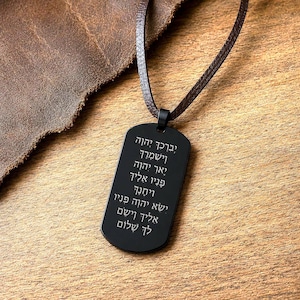 Könnte beinhalten: Schwarzer Dog-Tag-Anhänger mit hebräischem Text, an einer braunen Lederschnur befestigt. Der Anhänger ist rechteckig mit abgerundeten Ecken und gravierten weißen Buchstaben. Der Hintergrund ist eine Holzoberfläche.